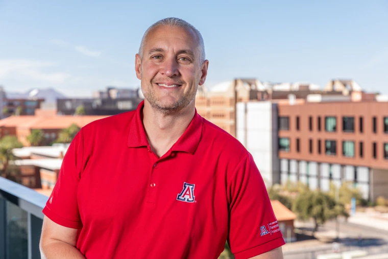 A photo of Denny Goforth who is on the Parent and Family Association Board, standing on a balcony with the Western Skyline behind him over looking the Unviersity of Arizona campus